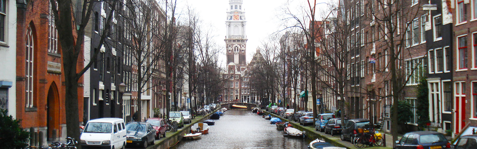 A canal with small boats is lined with tall and narrow houses. On the horizon is a bridge, and behind that is a tall church spire. 